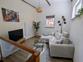 Ein modernes Wohnzimmer mit einem bequemen Sofa und einem Fernseher. Es gibt eine Pflanze und ein Kunstwerk an der Wand, sowie viel Tageslicht.