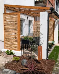 Ein gemütlicher Außenbereich mit Holzlatten und einem Tisch mit Stühlen. Eine Willkommens-Säule steht im Garten, umgeben von Pflanzen.