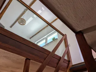 Ein moderner Raum mit einer Holztreppe und einer Glasbrüstung. An der Wand sind helle Fenster, die Tageslicht hereinlassen.