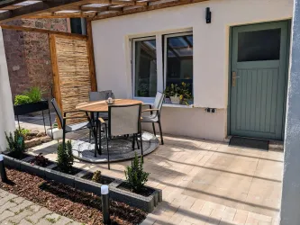 Ein gemütlicher Außenbereich mit einem runden Tisch und Stühlen. Die Terrasse ist hell und einladend, umgeben von Pflanzen.