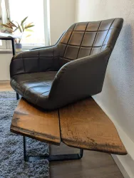 Ein moderner Stuhl mit einem grauen Kunstledersitz steht auf einer rustikalen Holzplatte. Der Stuhl hat eine ansprechende Form und ist in einem hellen Raum platziert.