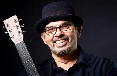 Ein lächelnder Mann mit einer Gitarre in der Hand trägt einen schwarzen Hut und eine Brille. Der Hintergrund ist dunkel und hebt seine positive Ausstrahlung hervor.