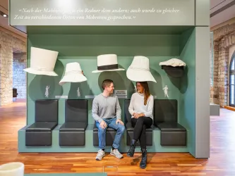 Eine Ausstellung mit großen Hüten und zwei Personen, die auf einer Bank sitzen. Im Hintergrund sind Zitate und eine moderne Wandgestaltung sichtbar.