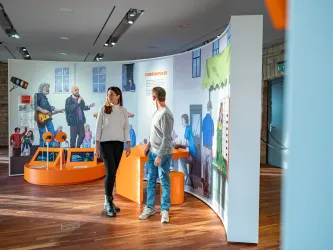 Zwei Personen stehen in einem modernen Ausstellungsraum und betrachten interaktive Displays. Im Hintergrund sind bunte Illustrationen und Ausstellungsstücke zu sehen.