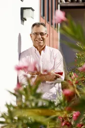 Ein fröhlicher Mann steht an einer Wand und lächelt. Im Vordergrund sind pinke Blumen zu sehen.