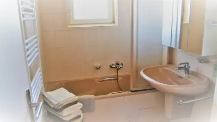 Ein Badezimmer mit einer Badewanne, einem Waschbecken und einem Spiegel. Die Wände sind hell gefliest und es liegen frische Handtücher bereit.