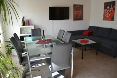 Ein modern eingerichtetes Wohnzimmer mit einem grauen Sofa und einem Glasstisch. Im Hintergrund befindet sich ein Fernseher und dekorative Bilder an der Wand.