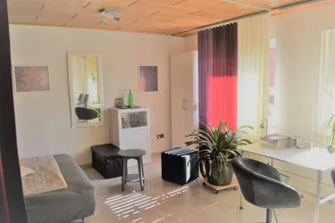 Ein modernes Zimmer mit einer Sitzgelegenheit und einem Schreibtisch. Große Fenster ermöglichen viel Licht und eine grüne Pflanze sorgt für eine gemütliche Atmosphäre.