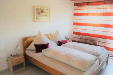 Ein gemütliches Schlafzimmer mit einem Doppelbett und zwei Kissen. Helle Wände und gestreifte Vorhänge sorgen für eine freundliche Atmosphäre.