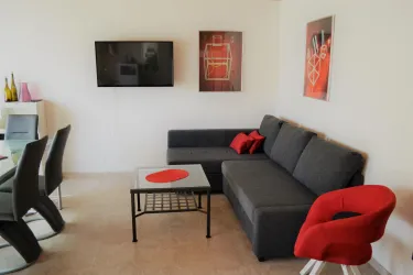 Ein modernes Wohnzimmer mit einem grauen Sofa und einem roten Stuhl. An den Wänden hängen Bilder, und ein Fernseher ist montiert.