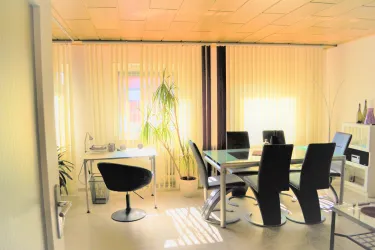 Ein modernes Büro mit einem großen Tisch und schwarzen Stühlen. Helle Fenster und Pflanzen sorgen für eine freundliche Atmosphäre.