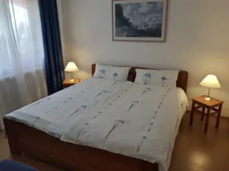Ein gemütliches Schlafzimmer mit einem großen Doppelbett und hellen Bettdecken. Zwei Nachttische mit Lampen und ein Fenster, das Tageslicht hereinlässt.