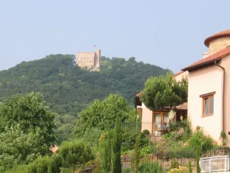 Eine malerische Landschaft mit einem Haus im Vordergrund und einer Burg auf einem Hügel im Hintergrund. Umgeben von üppigem Grün und Bäumen.