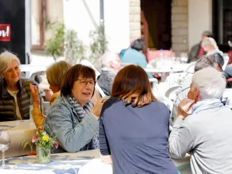 Eine Gruppe von Menschen sitzt draußen an Tischen und unterhält sich. Es scheint eine freundliche und gesellige Atmosphäre zu sein.