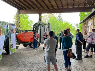 Eine Gruppe von Menschen hört einem Vortrag in einer Scheune zu. Im Hintergrund sind landwirtschaftliche Maschinen und Informationsplakate zu sehen.