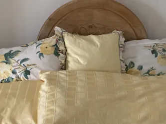 Ein gemütliches Bett mit gelben Kissen und einer blumigen Bettwäsche. Der Kopfteil ist aus Holz und hat ein einfaches, warmes Design.