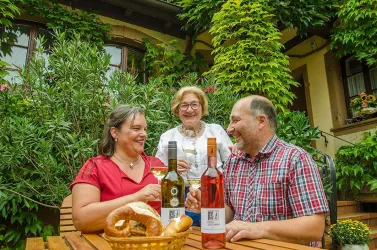 Eine fröhliche Gruppe von drei Personen genießt Wein an einem Tisch im Freien. Umgeben von Pflanzen und einer gemütlichen Atmosphäre feiern sie miteinander.