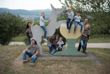 Eine Gruppe von Erwachsenen und Kindern posiert fröhlich um eine große Einhorn-Skulptur. Im Hintergrund sind sanfte Hügel und ein klarer Himmel sichtbar.