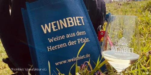 Eine blaue Tasche mit der Aufschrift "WEINBIET" steht neben einem Weinglas. Im Hintergrund sind grüne Wiesen zu sehen.