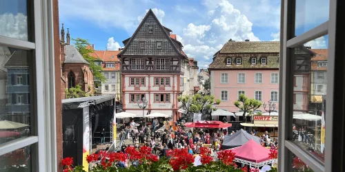 Ein belebter Marktplatz mit vielen Menschen und bunten Ständen. Historische Gebäude und blauer Himmel ergänzen die lebhafte Atmosphäre.