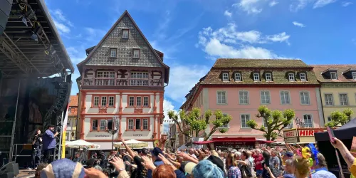 Ein belebter Platz mit Menschen, die ein Konzert genießen. Im Hintergrund sind schöne historische Gebäude und ein blauer Himmel zu sehen.