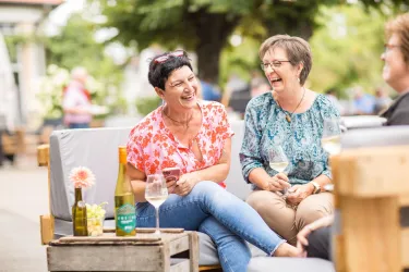 Zwei Frauen sitzen auf einer Couch und lachen herzlich. Auf dem Tisch stehen Gläser mit Weißwein und eine Flasche.