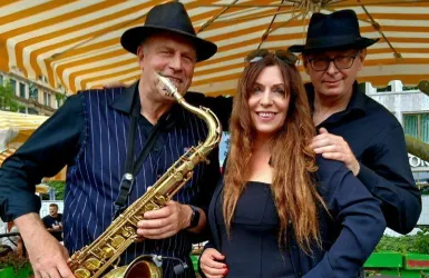 Drei Musiker stehen unter einem bunten Sonnenschirm. Ein Saxophonist, eine Sängerin und ein weiterer Mann posieren lächelnd für das Foto.