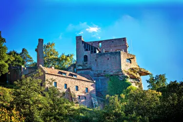 Eine alte Burg auf einem Hügel, umgeben von Bäumen und grünem Gelände. Der Himmel ist klar und blau.