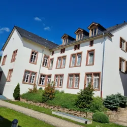 Ein modernes Gebäude mit einer hellen Fassade und charakteristischen Fenstern. Der Garten ist gepflegt und die Himmel ist blau mit wenigen Wolken.