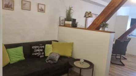 Ein gemütliches Wohnzimmer mit einem schwarzen Sofa und bunten Kissen. An der Wand hängen Bilder und Pflanzen sorgen für eine einladende Atmosphäre.
