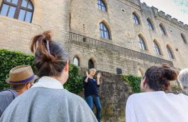 Eine Gruppe von Personen hört einer Rednerin zu, die vor einer historischen Burg spricht. Die Szenerie ist von grüner Vegetation umgeben und es ist ein klarer, sonniger Tag.