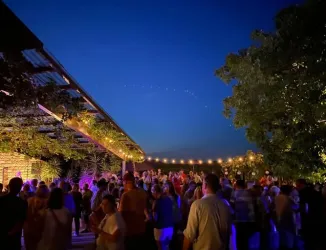 Eine lebhafte Feier im Freien bei Dämmerung. Viele Menschen tanzen und genießen die Atmosphäre unter Lichtern und Pflanzen.