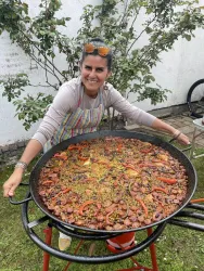 Eine Frau lächelt stolz und hält eine große Paella-Pfanne mit buntem Gemüse und Fleisch. Im Hintergrund sind Pflanzen und ein Fahrrad zu sehen.