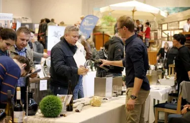 Eine Weinmesse mit verschiedenen Ständen und Besuchern, die Weine probieren. Mehrere Tische sind mit Flaschen und Gläsern dekoriert.