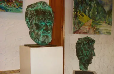 Zwei Skulpturen von Köpfen aus Bronze stehen in einer Galerie. Im Hintergrund sind bunte Gemälde zu sehen.