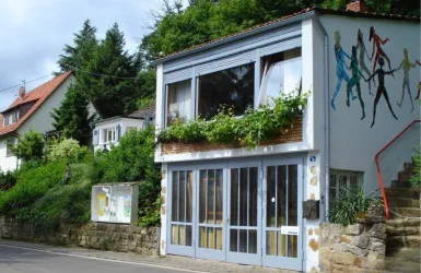Ein modernes Haus mit großen Fenstern und einem Blumenkasten. Im Hintergrund sind grüne Bäume und eine sanfte Hügelkette zu sehen.