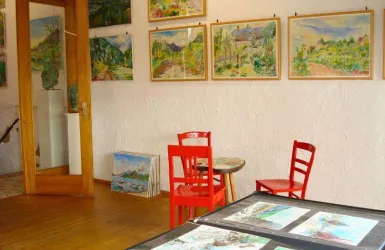 Einladende Galerie mit farbenfrohen Landschaftsgemälden an den Wänden. Rote Stühle und ein Tisch schaffen eine gemütliche Atmosphäre.