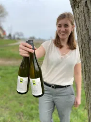 Eine Frau hält zwei Flaschen Wein in der Hand und lächelt. Im Hintergrund ist eine grüne Wiese und ein wolkiger Himmel zu sehen.