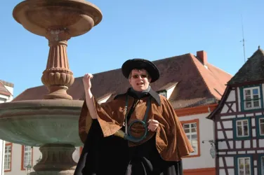 Eine Person in historischer Kleidung steht vor einem Brunnen und lächelt. Im Hintergrund sind traditionelle Gebäude mit roten Dächern zu sehen.