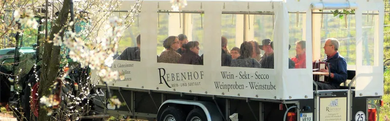 Ein Wagentrailer mit Passagieren, die an einer Weintour teilnehmen. Im Hintergrund sind blühende Pflanzen zu sehen.