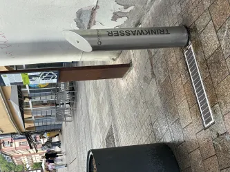 Eine Stadtstraße mit einem modernen Trinkwasserbrunnen. Im Hintergrund sind Tische und Stühle eines Cafés zu sehen.