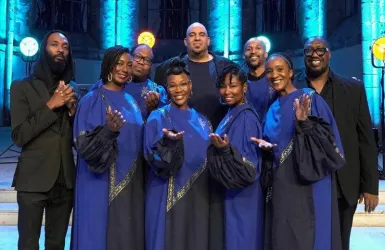 Eine Gruppe von Sängern in blauen Gewändern steht lächelnd zusammen. Im Hintergrund sind farbige Lichter in einer Kirche zu sehen.