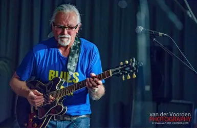 Ein Musiker spielt Gitarre auf der Bühne. Er trägt ein blaues T-Shirt mit der Aufschrift "BLUES" und hat weiße Haare.