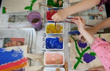 Kinder malen mit Pinsel auf Stofftaschen. Es gibt verschiedene Farben auf einem Tisch mit Zeitungen als Unterlage.