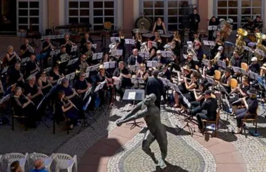 Ein Blasorchester spielt im Freien, umgeben von Zuschauern. Im Vordergrund steht eine Statue, die die Szenerie ergänzt.