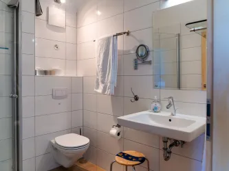 Ein modernes Badezimmer mit weißen Fliesen und einer Dusche. Es gibt ein Waschbecken, eine Toilette und einen kleinen Hocker.