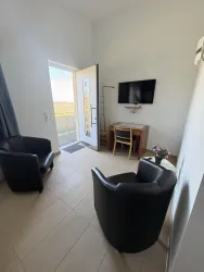 Ein helles Zimmer mit zwei schwarzen Sesseln, einem Tisch und einem Fernseher. Die Tür führt nach draußen und bietet einen Blick auf die Umgebung.