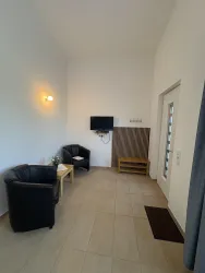 Ein helles Zimmer mit zwei schwarzen Sesseln und einem kleinen Tisch. An der Wand hängt ein Fernseher und es gibt einen Eingang mit einer Glastür.