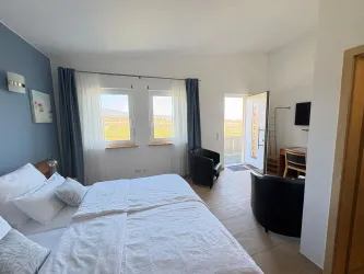 Ein helles Zimmer mit einem großen Bett und modernen Möbeln. Es gibt Fenster mit Aussicht auf die Landschaft und einen Zugang zur Terrasse.