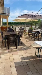 Ein gemütlicher Außenbereich mit Tischen und Stühlen unter Sonnenschirmen. Der Boden ist aus Stein und im Hintergrund sind grüne Pflanzen zu sehen.